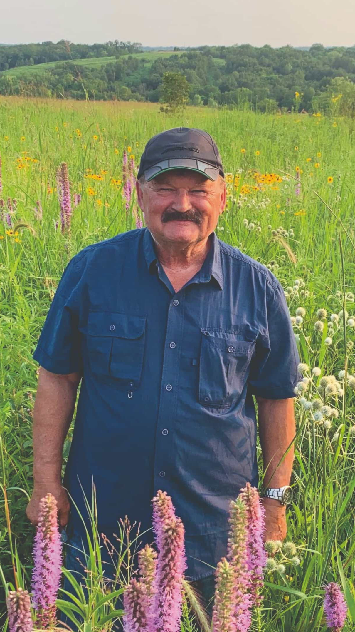 A profile photo of Rudi Roeslein smiling while standing outside in a prairie with long grass and flowers in the background.